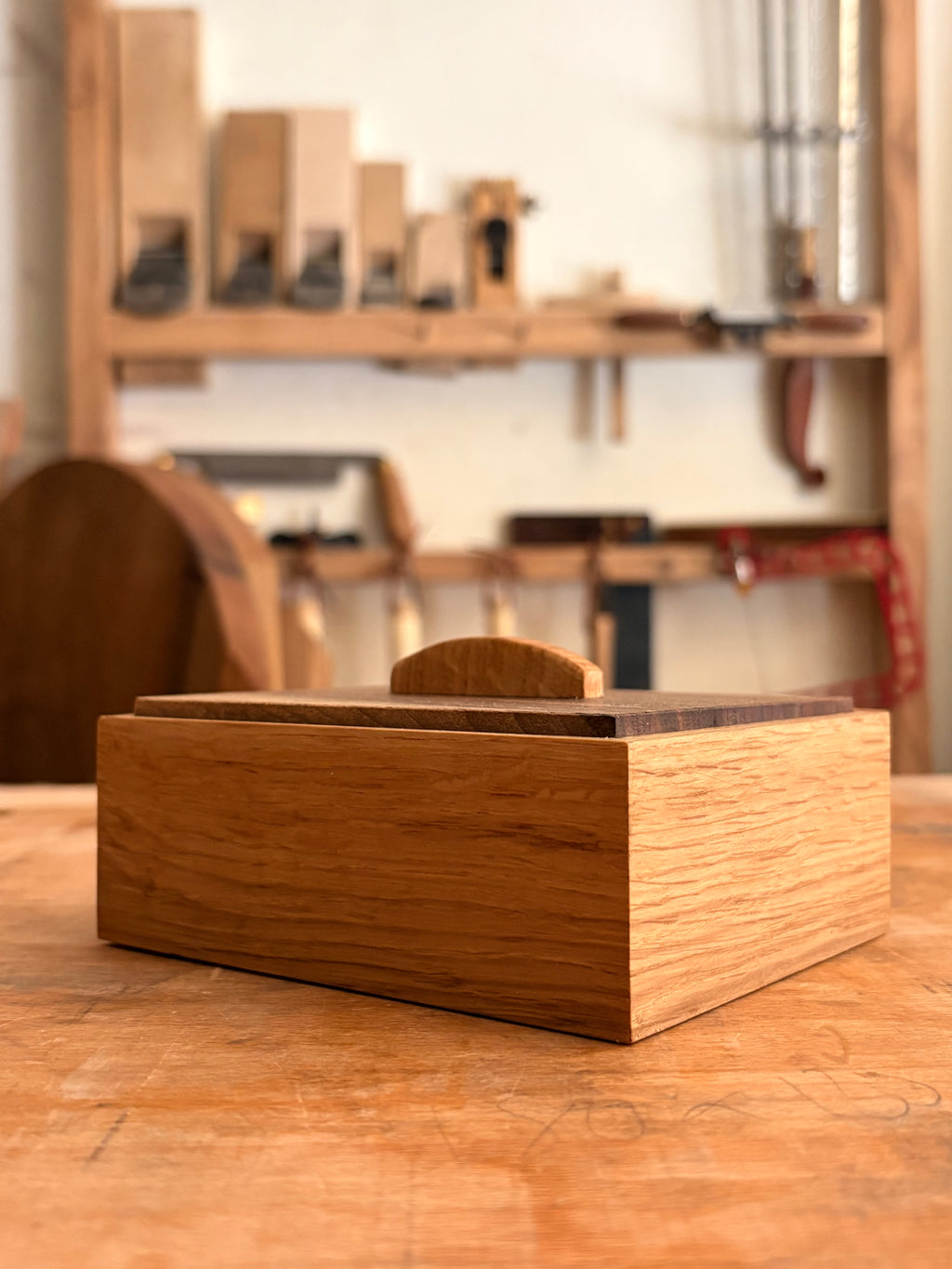 A rectangular wooden keepsake box with a push fit lid and a small arched pull, shown on a wooden workbench in a blurred woodworking workshop.