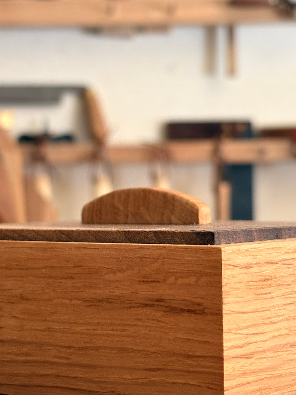 A close-up of a wooden box or furniture piece with a light brown wood grain front, a dark top surface, and a small matching wooden handle, photographed in a softly blurred workshop setting.