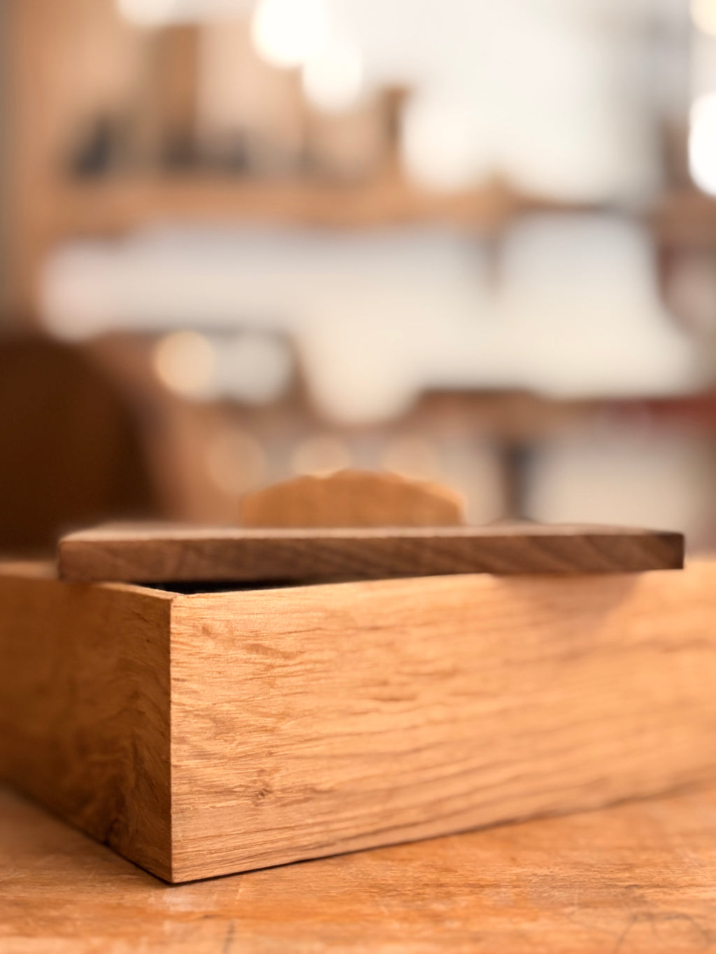 A close-up of a light brown wooden box with a matching lid resting on top, set on a wooden surface with a softly blurred background.