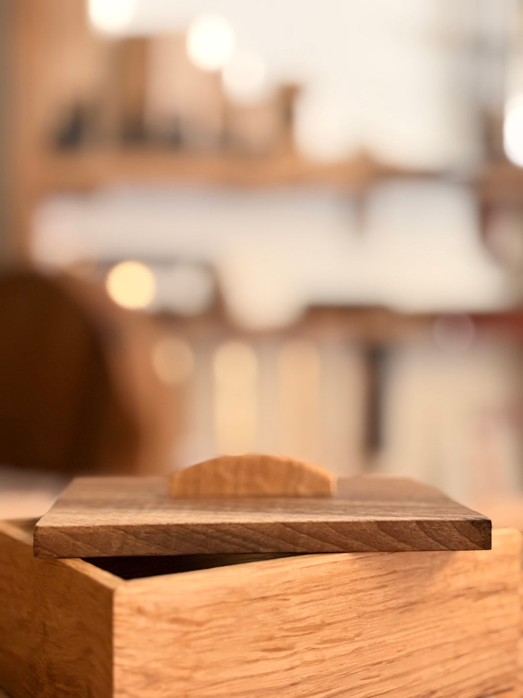 A close-up of a wooden storage box with a matching lid featuring a small arched handle, set against a heavily blurred warm-toned indoor background.